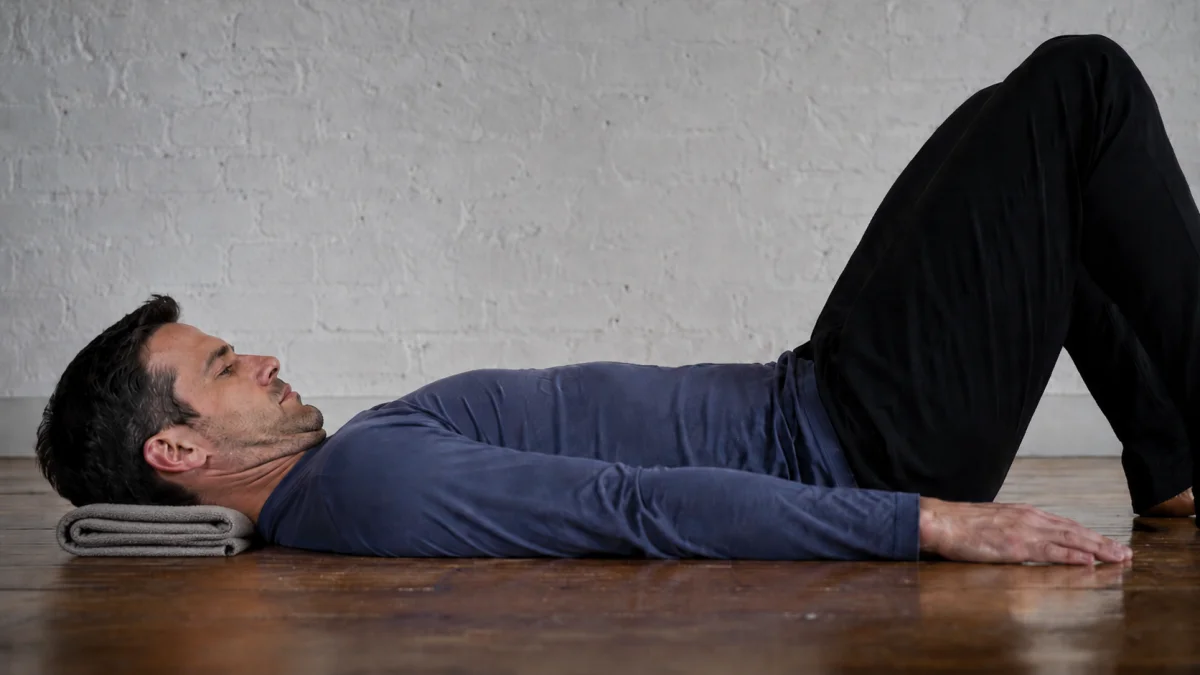 A man lying on his back on a wooden floor with his knees bent and his head resting on a folded towel, drawing his chin gently back toward his throat so the back of his neck lengthens toward the floor, with his head staying down rather than lifting